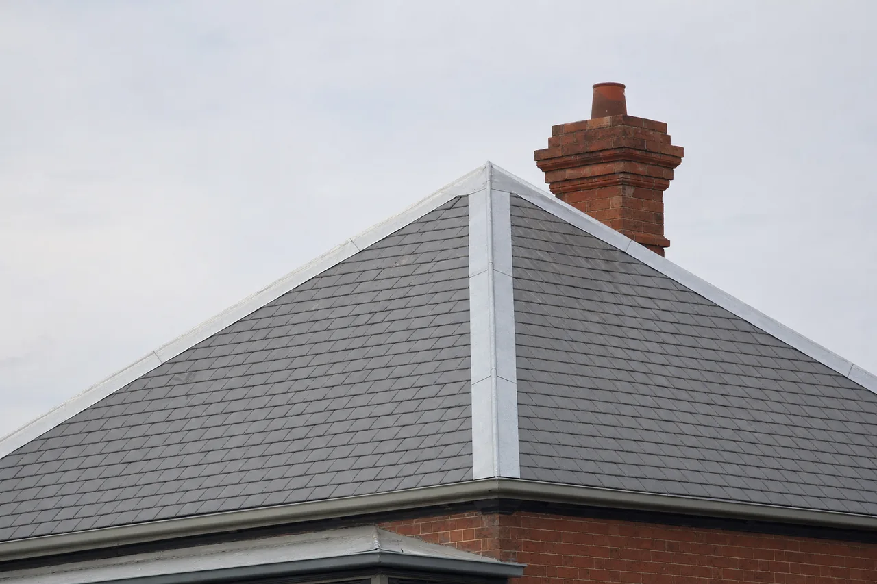 A hipped slate roof with lead-capped hips and a brick chimney against a pale sky — slate roofing, Ballarat. Illustrative example.